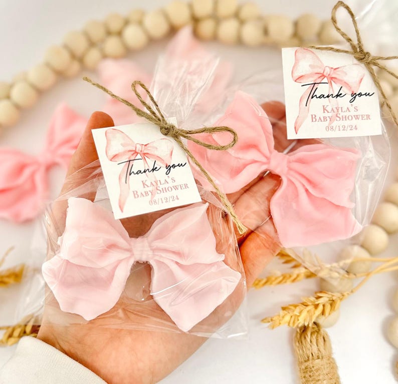 May include: Pink bow-shaped favors in clear cellophane bags, tied with twine and small tags. The tags read "Thank you, Kayla's Baby Shower 08/12/24". The bows are a soft, pastel pink color, perfect for a baby shower.