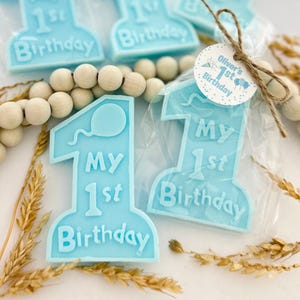 May include: Light blue number one shaped party favors with the text "My 1st Birthday" and a balloon graphic. Each favor is individually wrapped in clear cellophane. A small tag reads "Oliver's 1st Birthday".