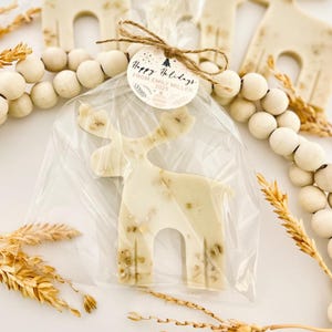 May include: A white reindeer-shaped treat with visible inclusions, packaged in clear cellophane and tied with twine. A round tag reads "Happy Holidays From Emily Miller 2025". The festive treat is surrounded by decorative wooden beads and wheat stalks.