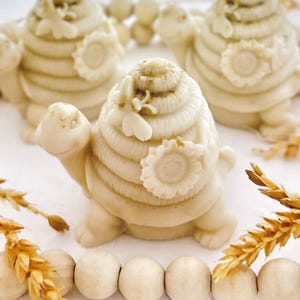 May include: Ivory-colored turtle figurines with beehive-shaped shells, adorned with sunflower and bee details. The figurines are arranged with wheat stalks and small, round objects, creating a nature-inspired theme.