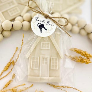 May include: A white house-shaped favor in a clear cellophane bag, tied with twine and a "Thank You" tag with keys. The house has windows, a door, and a chimney. Decorative wooden beads and wheat stalks are in the background.