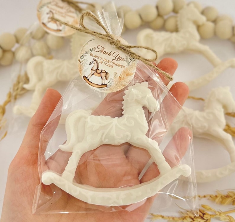 Puede incluir: Un favor de caballo de balanc&iacute;n blanco en una bolsa de celof&aacute;n transparente con un lazo de cuerda marr&oacute;n y una etiqueta de agradecimiento que dice "Gracias Blake's Baby Shower 09.17.2023"