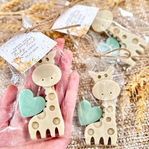 May include: Thank you for showering Baby Noah with love 05.17.2024.  A set of two favors in clear cellophane bags. Each bag contains a small green heart-shaped soap and a beige giraffe-shaped soap. The giraffe soap has a brown spot pattern.