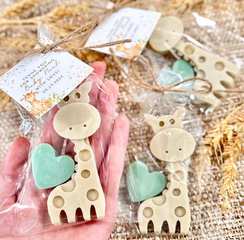 May include: Thank you for showering Baby Noah with love 05.17.2024.  A set of two favors in clear cellophane bags. Each bag contains a small green heart-shaped soap and a beige giraffe-shaped soap. The giraffe soap has a brown spot pattern.