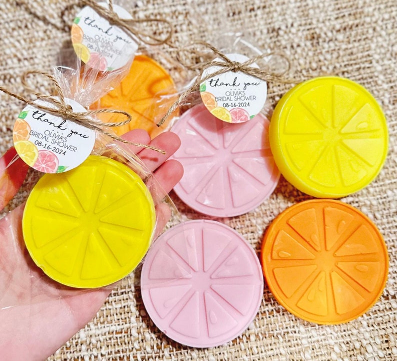 May include: Assortment of six round lemon-shaped soaps in yellow, orange, and pink. Each soap has a raised design of a lemon slice. The soaps are packaged in clear cellophane bags with a small tag that says "Thank you Olivia's Bridal Shower 08-16-2024".