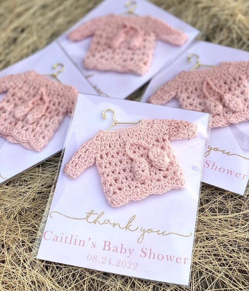 May include: Pink crocheted baby sweater favors with a gold hanger and a thank you tag that reads "Caitlin's Baby Shower 08.24.2022"