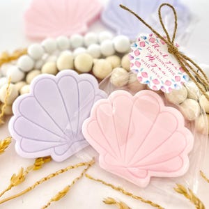May include: Two seashell-shaped favors, one pink and one lavender, are visible in a clear cellophane bag. The bag is tied with twine and a tag that reads "Thank you for showering Megan with love!" Wooden beads and dried wheat stalks are also in the image.