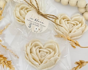 Puede incluir: Una pastilla de jabón en forma de corazón, blanca y floral, en una bolsa de plástico transparente con un lazo de cuerda marrón. Una pegatina redonda, blanca, con temática floral con el texto "LOVE in bloom" y "Ashley's Bridal Shower 09.12.2023" está pegada a la bolsa.