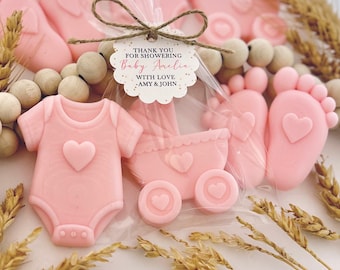 May include: Light pink baby-themed soaps shaped like onesies, strollers, and feet, packaged in clear plastic with a scalloped tag reading: THANK YOU FOR SHOWERING Baby Amelia WITH LOVE - AMY & JOHN, tied with twine and surrounded by wooden beads.
