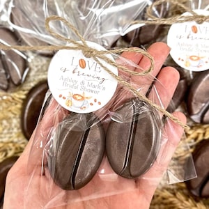 May include: Two brown chocolate coffee bean shaped favors wrapped in clear cellophane bags with a brown twine bow and a round tag that reads "LOVE is brewing Ashley & Mark's Bridal Shower".