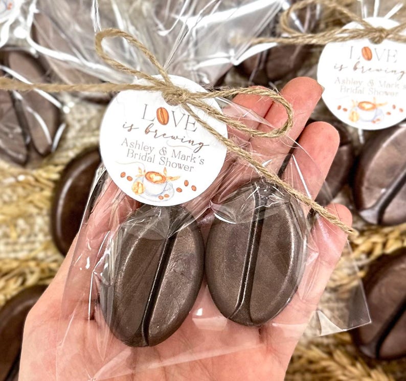 May include: Two brown chocolate coffee bean shaped favors wrapped in clear cellophane bags with a brown twine bow and a round tag that reads "LOVE is brewing Ashley & Mark's Bridal Shower".