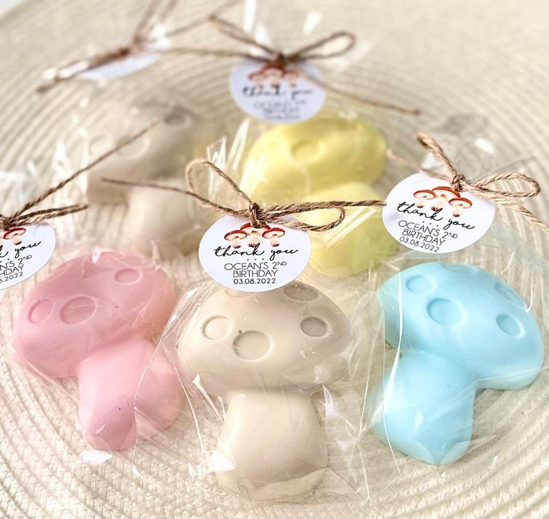 May include: Mushroom shaped soaps in pink, beige, yellow and blue, wrapped in cellophane bags with a brown twine bow. Each bag has a round white tag with the text "thank you OCEAN'S 2ND BIRTHDAY 03.08.2022" printed in black.