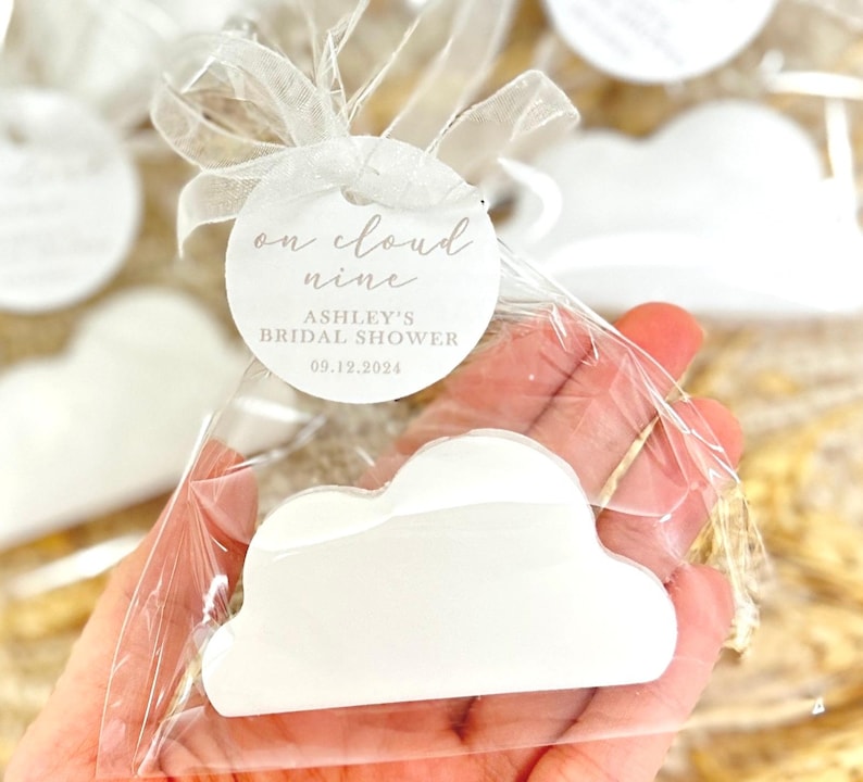 May include: White cloud-shaped favor with a round tag that says "on cloud nine" and "Ashley's Bridal Shower 09.12.2024".
