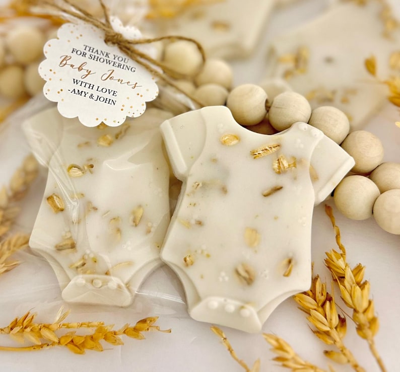 Op de afbeelding: Baby shower gunsten in de vorm van witte rompertjes, versierd met goudvlokken en haver. Elke gunst is verpakt in doorzichtig cellofaan en vastgebonden met touw en een label met de tekst "THANK YOU FOR SHOWERING Baby Jones WITH LOVE - AMY & JOHN."