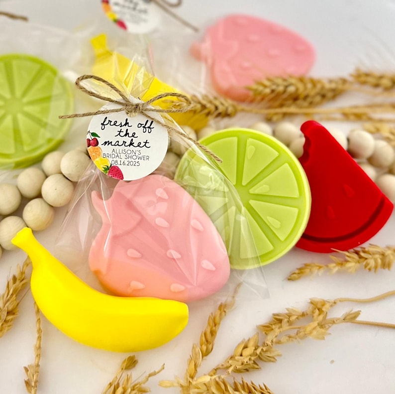May include: A set of five colorful fruit-shaped soaps in individual cellophane bags. The soaps are shaped like a banana, strawberry, lime, watermelon, and apple. The bags are tied with twine and a tag that reads "fresh off the market Allison's Bridal Shower 08.16.2025".