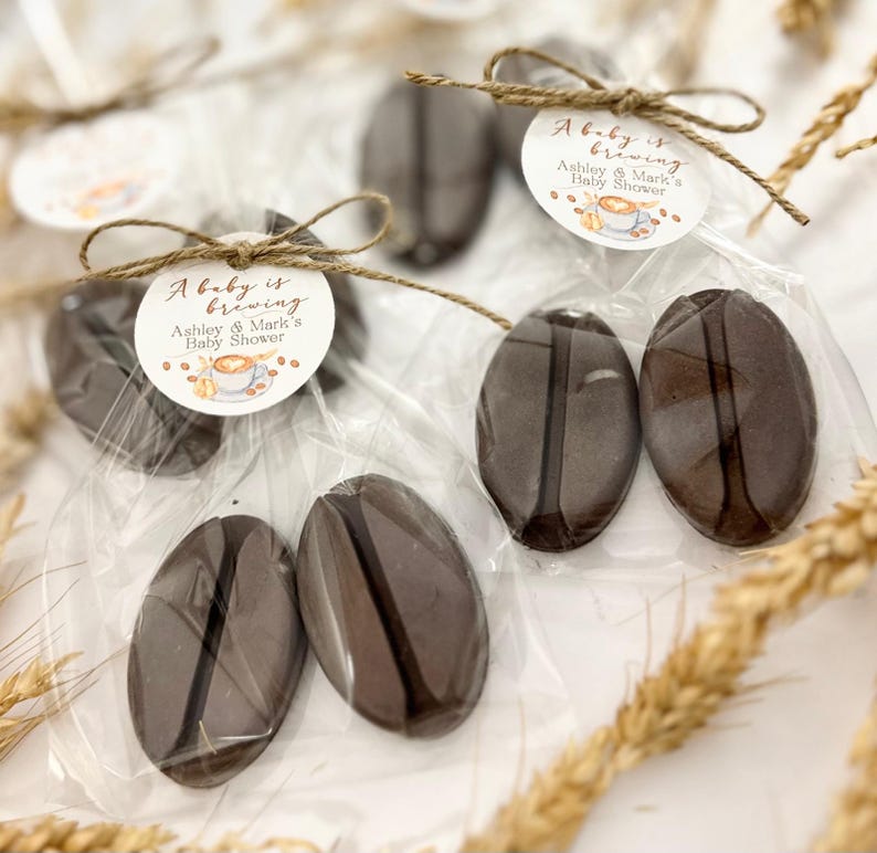 May include: Coffee bean-shaped favors in clear cellophane bags, tied with twine and a circular tag. The tag reads "A baby is brewing" with the names "Ashley & Mark's Baby Shower" and a coffee cup illustration. The favors are a dark brown color.