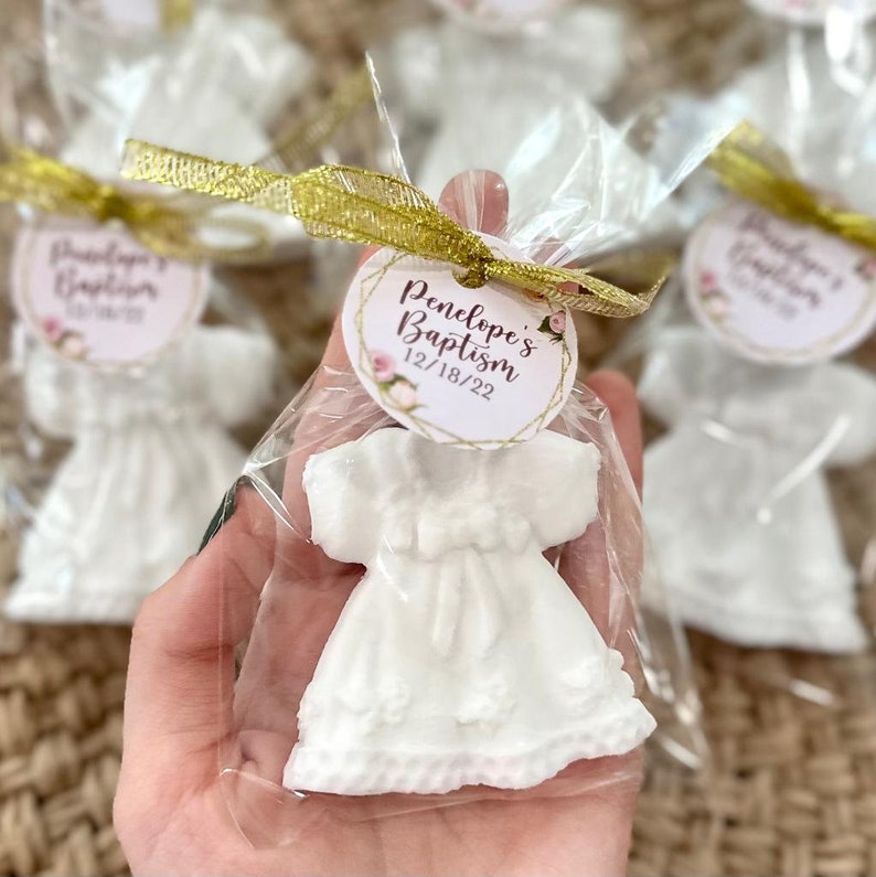 May include: White ceramic dress-shaped favour with a gold ribbon and a white tag with the text "Penelope's Baptism 12/18/22" in brown lettering.