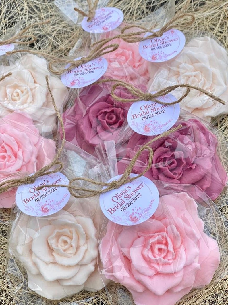 Puede incluir: Un conjunto de seis favores para una despedida de soltera, cada uno con una rosa de jab&oacute;n hecha a mano en una bolsa de pl&aacute;stico transparente atada con una cuerda. Las rosas de jab&oacute;n son en tonos de rosa y blanco, y cada bolsa tiene una etiqueta redonda con el texto "Olivia's Bridal Shower 08/22/2022".