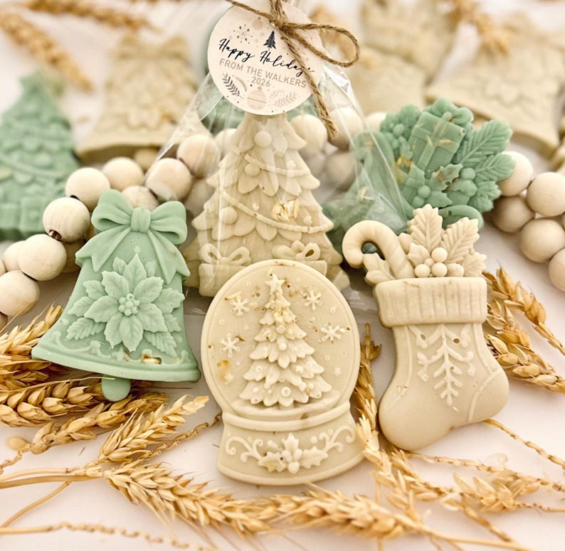 May include: A collection of holiday-themed wax melts in shapes of a bell, snow globe, and stocking, colored in sage green and cream. A clear bag contains a tree-shaped melt with a tag reading Happy Holidays FROM THE WALKERS 2026.
