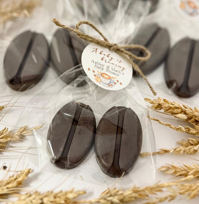 May include: A clear cellophane bag contains several dark brown, coffee bean-shaped treats. A small, round tag tied with twine reads "A baby is brewing" and "Ashley & Mark's Baby Shower". The treats are arranged with decorative wheat stalks.