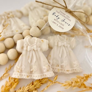 May include: Two ivory-colored baby dress-shaped soaps are displayed in a clear cellophane bag, tied with twine and a tag that reads "Baby Jones". The soaps have bow details and floral accents. Wooden beads and wheat stalks are also visible.