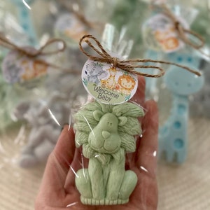 Puede incluir: Una pastilla de jab&oacute;n verde en forma de le&oacute;n con un lazo de cuerda marr&oacute;n y una etiqueta redonda que dice "Bienvenido Beb&eacute;" con un elefante, una jirafa y un le&oacute;n de dibujos animados.