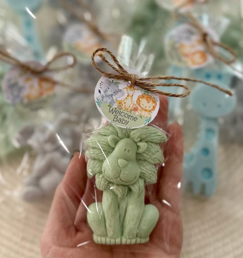 Puede incluir: Una pastilla de jab&oacute;n verde en forma de le&oacute;n con un lazo de cuerda marr&oacute;n y una etiqueta redonda que dice "Bienvenido Beb&eacute;" con un elefante, una jirafa y un le&oacute;n de dibujos animados.