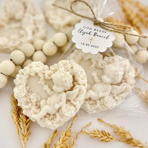May include: Ivory-colored heart-shaped soaps with a cherub and rose design. One soap is packaged in clear cellophane with a tag that reads "GOD BLESS Elijah Daniel" and the date 12.20.2025. Decorative elements include a wooden bead garland and wheat stalks.
