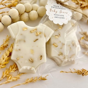 Op de afbeelding: Ivoor kleurige zeepjes in de vorm van een babyromper, versierd met gouden vlokken. Verpakt in transparant cellofaan. Een label leest: "Thank you for showering Baby Jones with love - Amy & John."