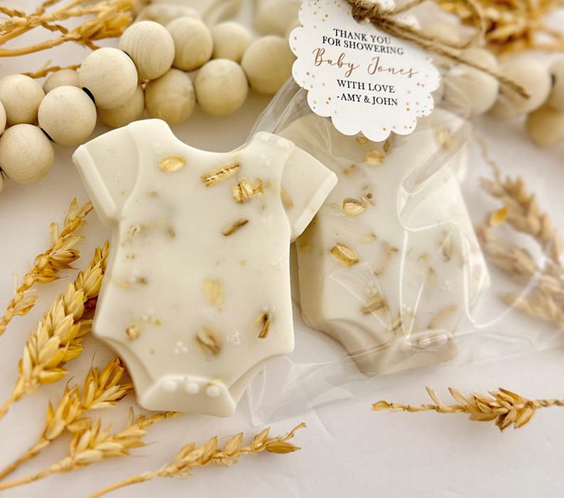 Op de afbeelding: Ivoor kleurige zeepjes in de vorm van een babyromper, versierd met gouden vlokken. Verpakt in transparant cellofaan. Een label leest: "Thank you for showering Baby Jones with love - Amy & John."