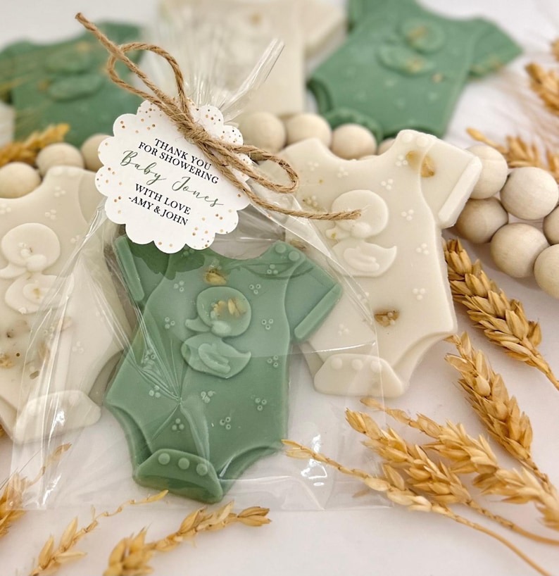 May include: Baby shower favors shaped like baby onesies in sage green and off-white, with a duck design. Each onesie is individually wrapped and tied with twine and a tag that reads "Thank you for showering Baby Jones with love - Amy & John."