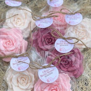 Puede incluir: Un conjunto de cinco jabones en forma de rosa rosa, blanco y rubor, envueltos en bolsas de celof&aacute;n transparentes con cordel y una etiqueta personalizada. La etiqueta dice "Olivia's Bridal Shower 08/22/2022".