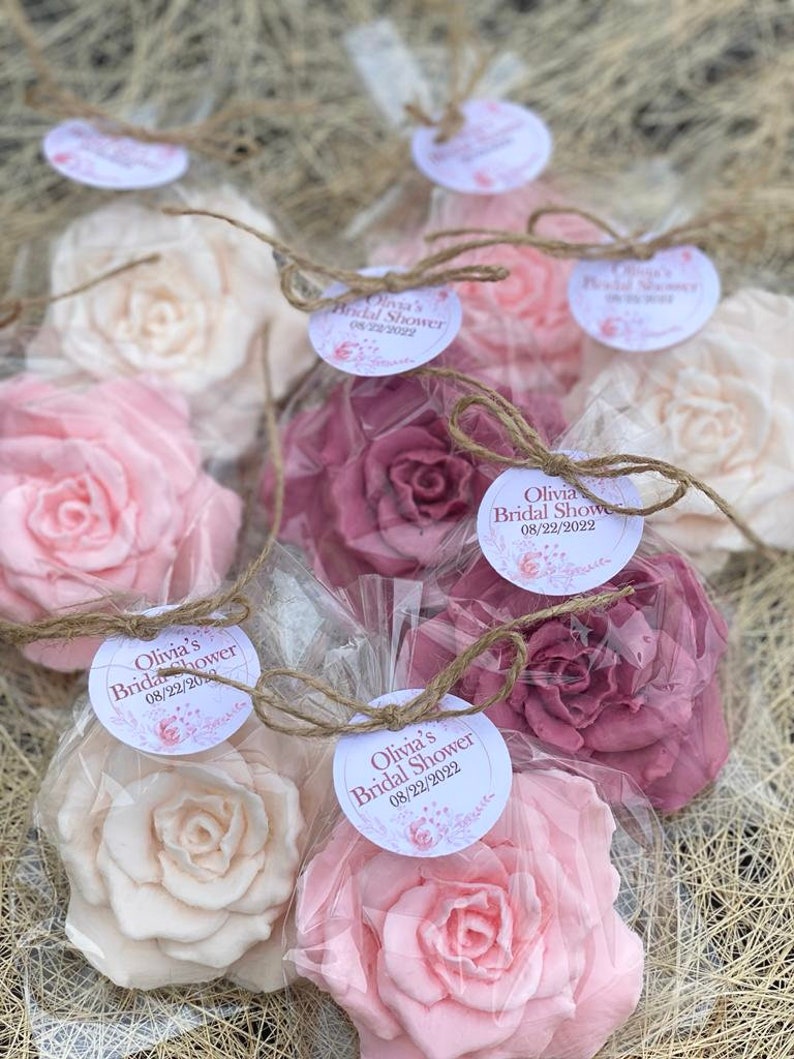 Puede incluir: Un conjunto de cinco jabones en forma de rosa rosa, blanco y rubor, envueltos en bolsas de celof&aacute;n transparentes con cordel y una etiqueta personalizada. La etiqueta dice "Olivia's Bridal Shower 08/22/2022".