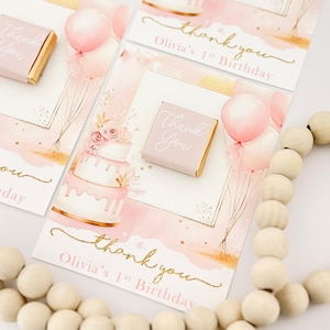 Puede incluir: Tarjetas de agradecimiento rosas y doradas con un pastel de acuarela y globos. Las tarjetas dicen "Gracias" y "Olivia's 1st Birthday".