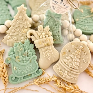 May include: A collection of decorative wax melts in cream and sage green, shaped like Christmas trees, stockings, sleighs, and snow globes. One item is in a clear bag with a tag reading Happy Holidays FROM THE WALKERS 2026. The items feature intricate embossed details like snowflakes, gifts, and holly.