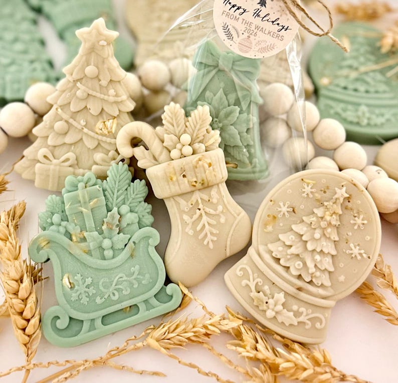 May include: A collection of decorative wax melts in cream and sage green, shaped like Christmas trees, stockings, sleighs, and snow globes. One item is in a clear bag with a tag reading Happy Holidays FROM THE WALKERS 2026. The items feature intricate embossed details like snowflakes, gifts, and holly.