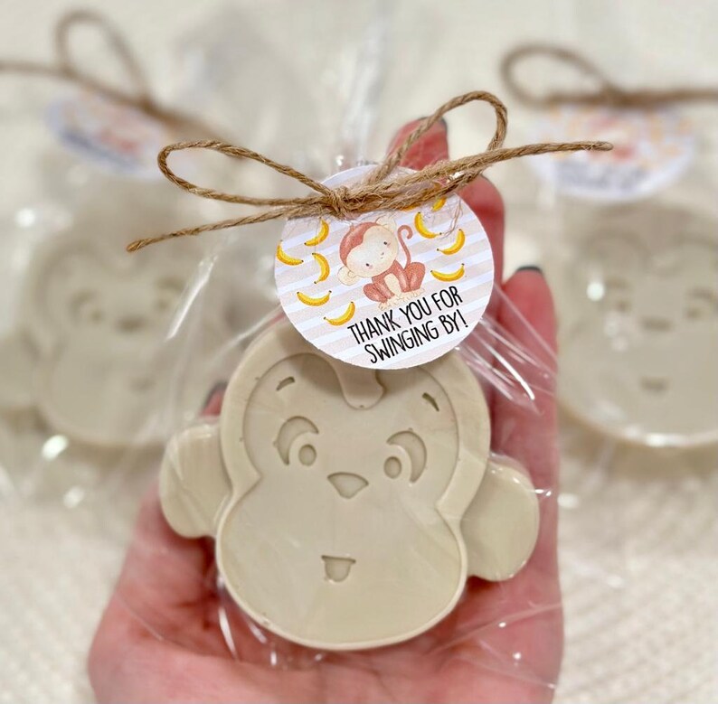 May include: A small, beige monkey-shaped soap bar with a cute face is wrapped in clear cellophane and tied with twine. A round tag with a monkey and bananas is attached to the twine. The tag reads "Thank you for swinging by!"