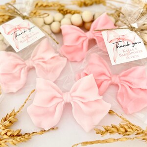 May include: Several light pink fabric bows in clear cellophane bags, tied with twine. Each bag has a tag with a watercolor bow illustration and the text "Thank you, Kayla's Baby Shower 08/12/24". Decorative wheat stalks and wooden beads are in the background.