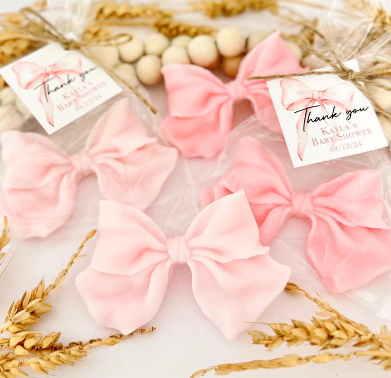 May include: Several light pink fabric bows in clear cellophane bags, tied with twine. Each bag has a tag with a watercolor bow illustration and the text "Thank you, Kayla's Baby Shower 08/12/24". Decorative wheat stalks and wooden beads are in the background.