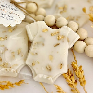Op de afbeelding: Baby shower gunsten in de vorm van een witte romper, versierd met goudvlokken en haver. Het label luidt "THANK YOU FOR SHOWERING Baby Jones WITH LOVE - AMY & JOHN."