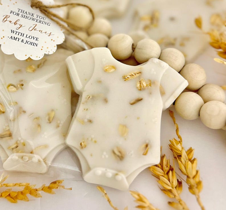 Op de afbeelding: Baby shower gunsten in de vorm van een witte romper, versierd met goudvlokken en haver. Het label luidt "THANK YOU FOR SHOWERING Baby Jones WITH LOVE - AMY & JOHN."