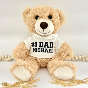 May include: A beige teddy bear wearing a white hooded shirt with the text "#1 DAD MICHAEL" printed in black.