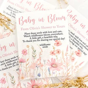May include: Pink and white floral design with the text "Baby in Bloom" and "From Olivia's Shower to Yours". The text continues with "Plant these seeds with love and care, Watch wildflowers bloom everywhere. A little gift, a heartfelt way, To thank you for sharing our special day!" and "wildflower seeds".