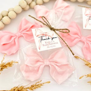 May include: Pink fabric bows packaged in clear plastic, tied with twine, and labeled with a "Thank you" tag for Kayla's Baby Shower on 08/12/24. The bows are a soft, pastel pink color, and are arranged on a white surface.
