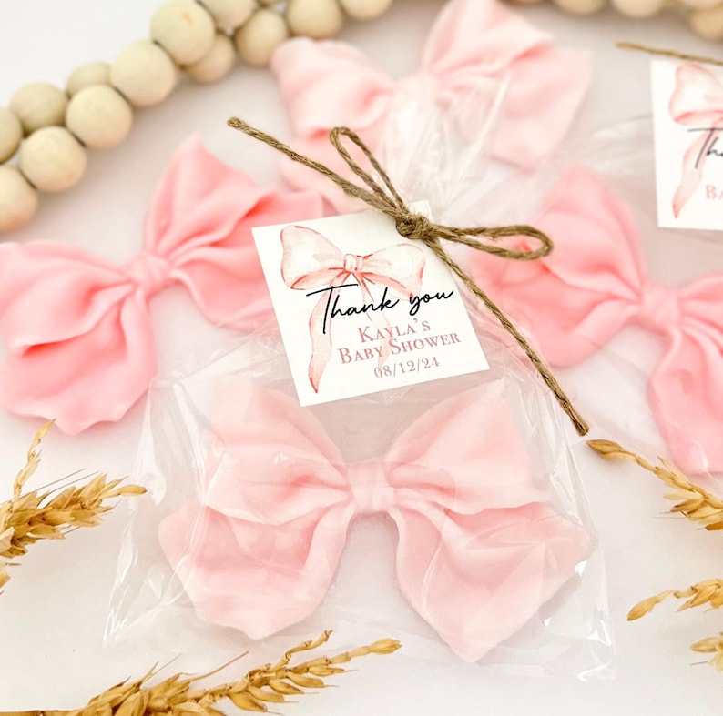 May include: Pink fabric bows packaged in clear plastic, tied with twine, and labeled with a "Thank you" tag for Kayla's Baby Shower on 08/12/24. The bows are a soft, pastel pink color, and are arranged on a white surface.