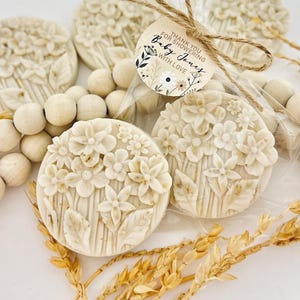 May include: Off-white, floral-shaped soaps with detailed flower and leaf designs. Some soaps are packaged with a tag that reads "Thank you for showering Baby Jones with love." The soaps are arranged with wooden beads and dried wheat.