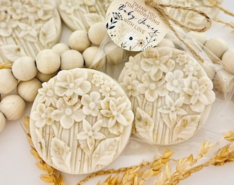 May include: Off-white, floral-shaped soaps with detailed flower and leaf designs. Some soaps are packaged with a tag that reads "Thank you for showering Baby Jones with love." The soaps are arranged with wooden beads and dried wheat.