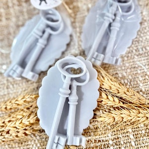 May include: Two sets of antique-style keys in a light gray oval frame. The keys are made of a smooth, light gray material and have intricate details. The frames are decorated with a scalloped edge.