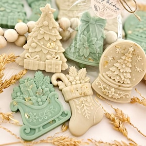 May include: A collection of decorative holiday-themed wax melts in sage green and cream colors. Shapes include a Christmas tree with gifts, a bell with a poinsettia, a snow globe with a tree, a sleigh filled with presents, and a stocking with holly.