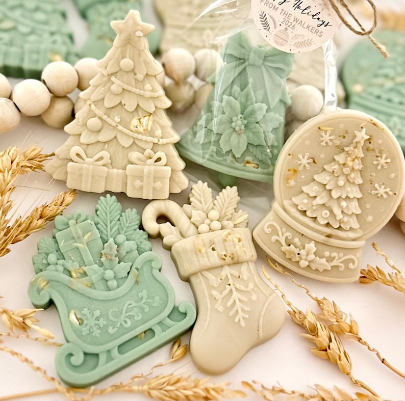 May include: A collection of decorative holiday-themed wax melts in sage green and cream colors. Shapes include a Christmas tree with gifts, a bell with a poinsettia, a snow globe with a tree, a sleigh filled with presents, and a stocking with holly.